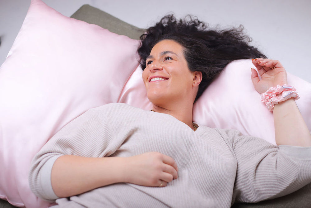Lady wearing two pink scrunchies on left wrist and lying on two pink silk pillowcases smiling